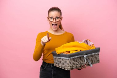 Pembe arka planda izole edilmiş çamaşır sepeti tutan beyaz bir kadın şaşırmış ve ön tarafı işaret ediyor.