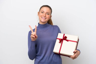 Beyaz arka planda izole edilmiş bir hediye tutan genç beyaz kadın gülümsüyor ve zafer işareti gösteriyor.