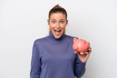 Beyaz arka planda, şaşırmış ve şok olmuş bir yüz ifadesiyle bir kumbarayı elinde tutan beyaz bir kadın.
