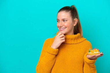 Kafkasyalı genç bir kadın elinde turtayla mavi arka planda izole edilmiş bir şekilde bir fikir üretiyor ve bir tarafa bakıyor.