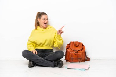 Beyaz arka planda beyaz bir zeminde oturan beyaz tenli genç bir kadın parmak göstererek bir ürün sunuyor.