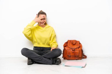 Beyaz arka planda, beyaz bir zeminde oturan beyaz tenli genç bir kadın elleri ile gözlerini kapatıyor ve gülümsüyor.