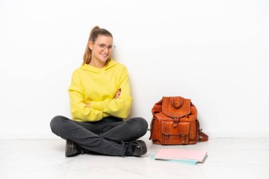 Beyaz arka planda beyaz bir zeminde oturan beyaz tenli genç bir kadın. Kolları bağlı ve ileriye bakıyor.