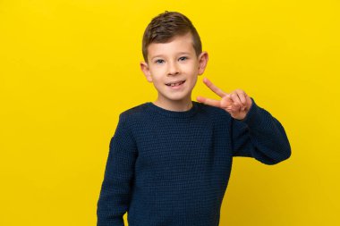Sarı arka planda izole edilmiş küçük beyaz çocuk gülümsüyor ve zafer işareti gösteriyor.