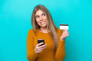Kafkasyalı genç bir kadın mavi arka planda izole edilmiş. Cep telefonuyla kredi kartıyla alışveriş yapıyor.