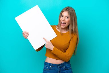 Kafkasyalı genç bir kadın mavi arka planda izole edilmiş elinde mutlu bir ifadeyle boş bir levha tutuyor ve onu işaret ediyor.