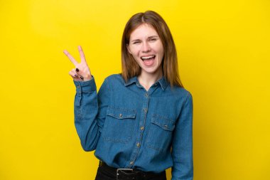 Sarı arka planda izole edilmiş genç İngiliz kadın gülümsüyor ve zafer işareti gösteriyor.