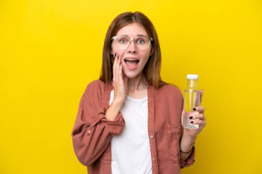 Sarı zemin üzerinde su şişesi olan genç İngiliz kadın şaşırmış ve şok olmuş yüz ifadesi ile izole edilmiş.