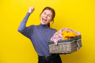 Sarı arka planda izole edilmiş çamaşır sepetini tutan genç İngiliz kadın zaferi kutluyor.