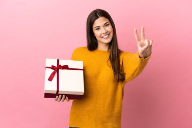 İzole edilmiş pembe arka planda bir hediye tutan Brezilyalı genç kız gülümsüyor ve zafer işareti gösteriyor.