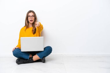 Yerde dizüstü bilgisayarlı beyaz bir kadın oturuyor. Beyaz arka planda izole edilmiş. Şaşırmış ve ön tarafı gösteriyor.