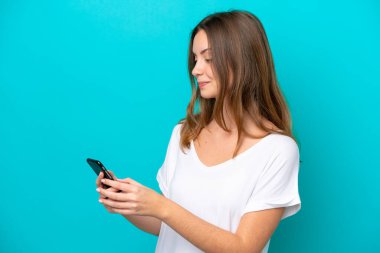 Mavi arka planda izole edilmiş genç beyaz kadın cep telefonuyla mesaj ya da e-posta gönderiyor.