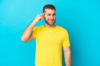 Genç, beyaz, mavi arka planda izole edilmiş bir adam deliliğin jestini yapıyor. Parmağını kafaya koyuyor.