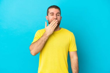 Mavi arka planda izole edilmiş yakışıklı beyaz adam esniyor ve ağzını eliyle açık tutuyor.