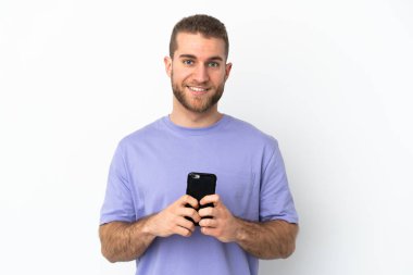 Beyaz arka planda izole edilmiş yakışıklı beyaz adam kameraya bakıyor ve cep telefonunu kullanarak gülümsüyor.