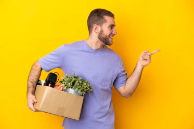 Genç Kafkasyalı, sarı arka planda izole edilmiş eşyalarla dolu bir kutuyu kaldırırken hareket ediyor. Parmakla yan tarafı gösteriyor ve bir ürün sunuyor.