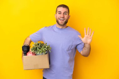 Genç Kafkasyalı, sarı arka planda izole edilmiş eşyalarla dolu bir kutuyu alırken hareket ediyor. Parmaklarıyla beşi sayıyor.