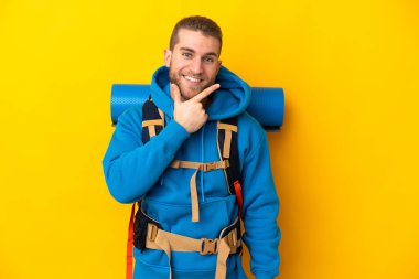 Sarı arka planda izole edilmiş büyük sırt çantalı beyaz bir dağcı. Mutlu ve gülümsüyor.