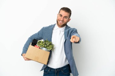 Beyaz arka planda izole edilmiş eşyalarla dolu bir kutuyu alıp mutlu bir ifadeyle öne doğru hareket eden genç beyaz tenli.