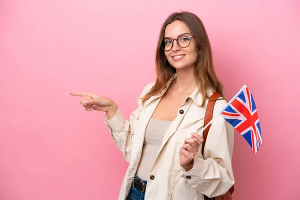 若い学生の白人女性側に指を指してピンクの背景に隔離された英国のフラグを保持 — ストック写真