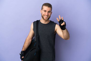 Spor çantalı genç adam beyaz arka planda izole edilmiş mutlu ve parmaklarıyla üçe kadar sayıyor.