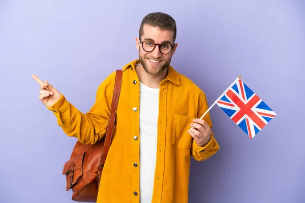 年轻的高加索人拿着一面英国国旗 用紫色背景隔离 手指指向侧面 — 图库照片