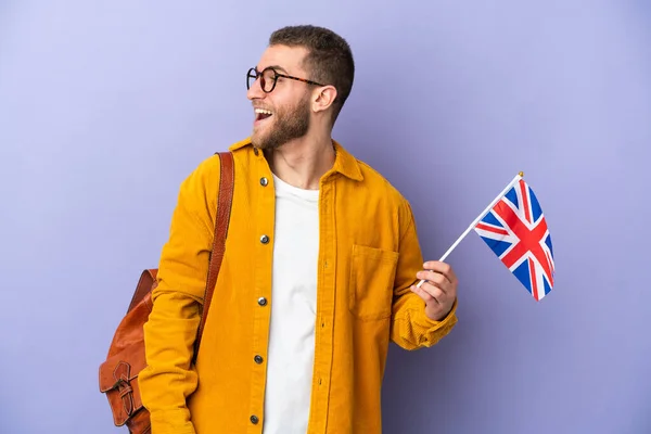 年轻的高加索男子举着一面英国国旗 被紫色背景隔离 在侧面笑着 — 图库照片