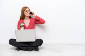 Mladá zrzka žena s notebookem sedí na podlaze izolované na bílém pozadí drží kávu odnést a mobilní