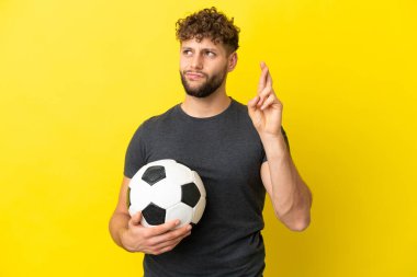 Yakışıklı genç futbolcu sarı arka planda izole edilmiş, parmaklarını çapraz yapıp en iyisini diliyor.