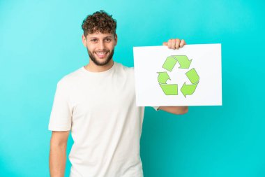 Mavi arka planda izole edilmiş yakışıklı beyaz bir adam elinde mutlu bir ifadeyle geri dönüşüm simgesiyle bir pankart tutuyor.