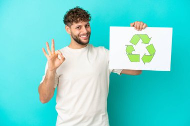 Mavi arka planda izole edilmiş yakışıklı beyaz adam elinde geri dönüşüm simgesiyle bir pankart tutuyor ve zaferi kutluyor.