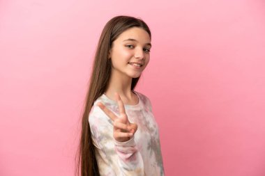 Küçük kız pembe arka planda gülümsüyor ve zafer işareti gösteriyor.