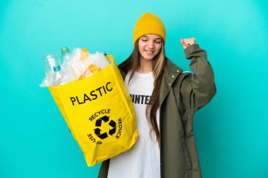 Bir çanta dolusu plastik şişeyi elinde tutan küçük kız izole edilmiş mavi arka planda zaferi kutlamak için geri dönüşüm yapıyor.