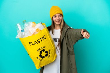 Bir çanta dolusu plastik şişeyi elinde tutan küçük kız izole edilmiş mavi arkaplanı geri dönüştürmek için baş parmak işareti yapıyor.