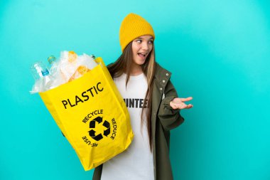 Bir çanta dolusu plastik şişeyi elinde tutan küçük kız izole edilmiş mavi arkaplanı süpriz bir ifadeyle geri dönüştürmek için yandan bakarken