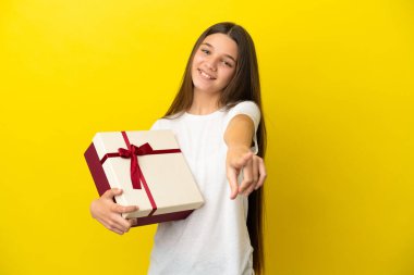 İzole edilmiş sarı arka planda elinde bir hediye tutan küçük kız mutlu bir ifadeyle öne bakıyor.