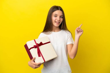Küçük kız sarı arka planda bir hediye tutuyor ve en iyinin işareti olarak parmağını kaldırıyor.