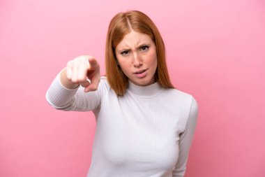 Genç kızıl saçlı kadın pembe arka planda izole edilmiş hayal kırıklığına uğramış ve ön tarafı işaret ediyor.