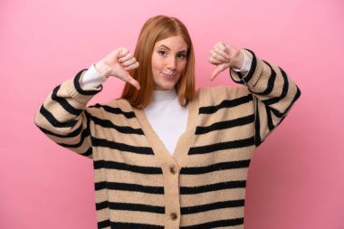 Pembe arka planda izole edilmiş genç kızıl saçlı kadın baş parmağını iki eliyle aşağı gösteriyor.