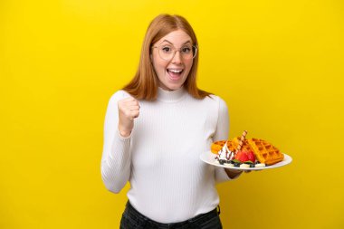 Sarı arka planda gözleme tutan genç kızıl saçlı kadın zaferi kutluyor.