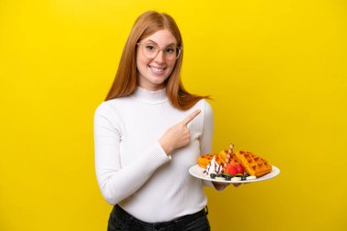 Sarı arka planda izole edilmiş waffle tutan genç kızıl saçlı kadın bir ürünü sunmak için yan tarafı işaret ediyor.