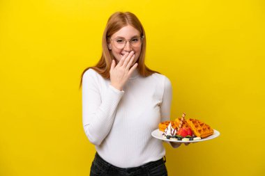 Sarı arka planda izole edilmiş waffle tutan genç kızıl saçlı kadın mutlu ve gülümsüyor.