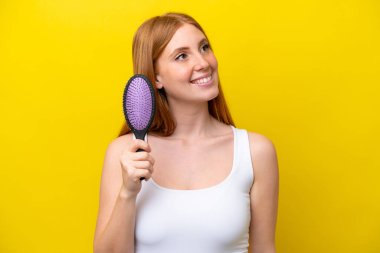 Kızıl saçlı genç bir kadın elinde saç fırçası ile beyaz arka planda izole edilmiş gülümserken yukarı bakıyor.