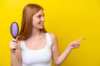 Kızıl saçlı genç bir kadın, elinde saç fırçası, beyaz arka planda izole edilmiş bir şekilde bir ürünü sunmak için yan tarafı işaret ediyor.