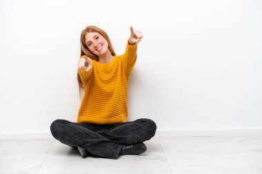 Genç kızıl saçlı kadın, beyaz arka planda izole bir şekilde yerde oturmuş gülümserken sana işaret ediyor.