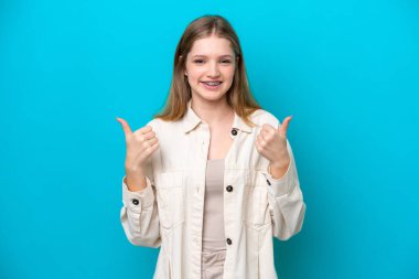 Genç Rus kız mavi arka planda izole edilmiş, başparmakları havada ve gülümsüyor.