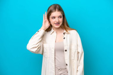 Genç Rus kız, mavi arka planda izole edilmiş bir şekilde elini kulağına koyarak bir şeyler dinliyor.