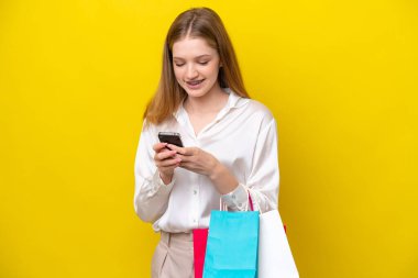 Sarı arka planda izole edilmiş genç bir Rus kız elinde alışveriş çantaları ve bir arkadaşına cep telefonuyla bir mesaj yazıyor.