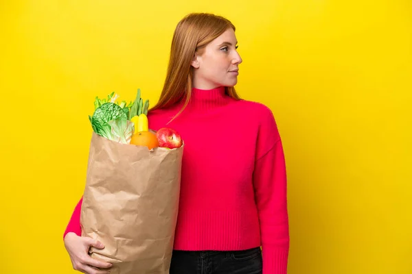 Молодая Рыжая Женщина Держит Сумку Продуктами Изолированы Желтом Фоне Глядя — стоковое фото