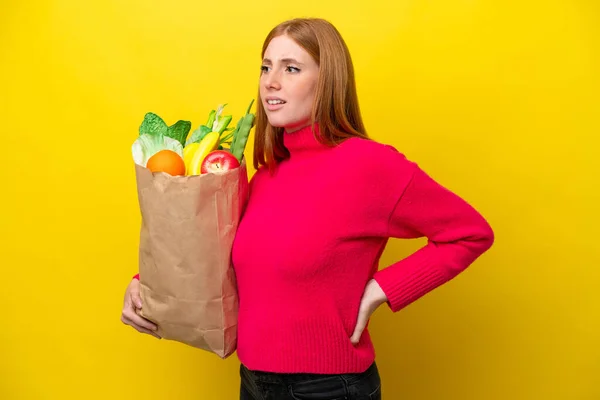 黄色の背景に孤立した食料品の買い物袋を持っている若い赤毛の女性は 努力をしたために腰痛に苦しんでいます — ストック写真